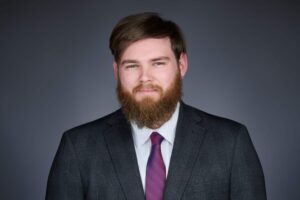 A bearded man in a dark plaid suit and purple tie is pictured against a gray background. He has short hair and is looking directly at the camera with a slight smile.