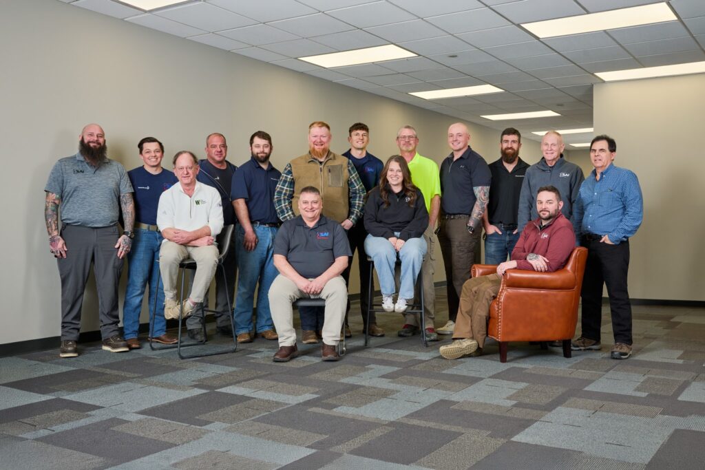 A group of 15 people, both men and women, pose together in an office setting. Some are seated while others stand behind them in casual attire, against a backdrop of a plain wall and tiled ceiling.