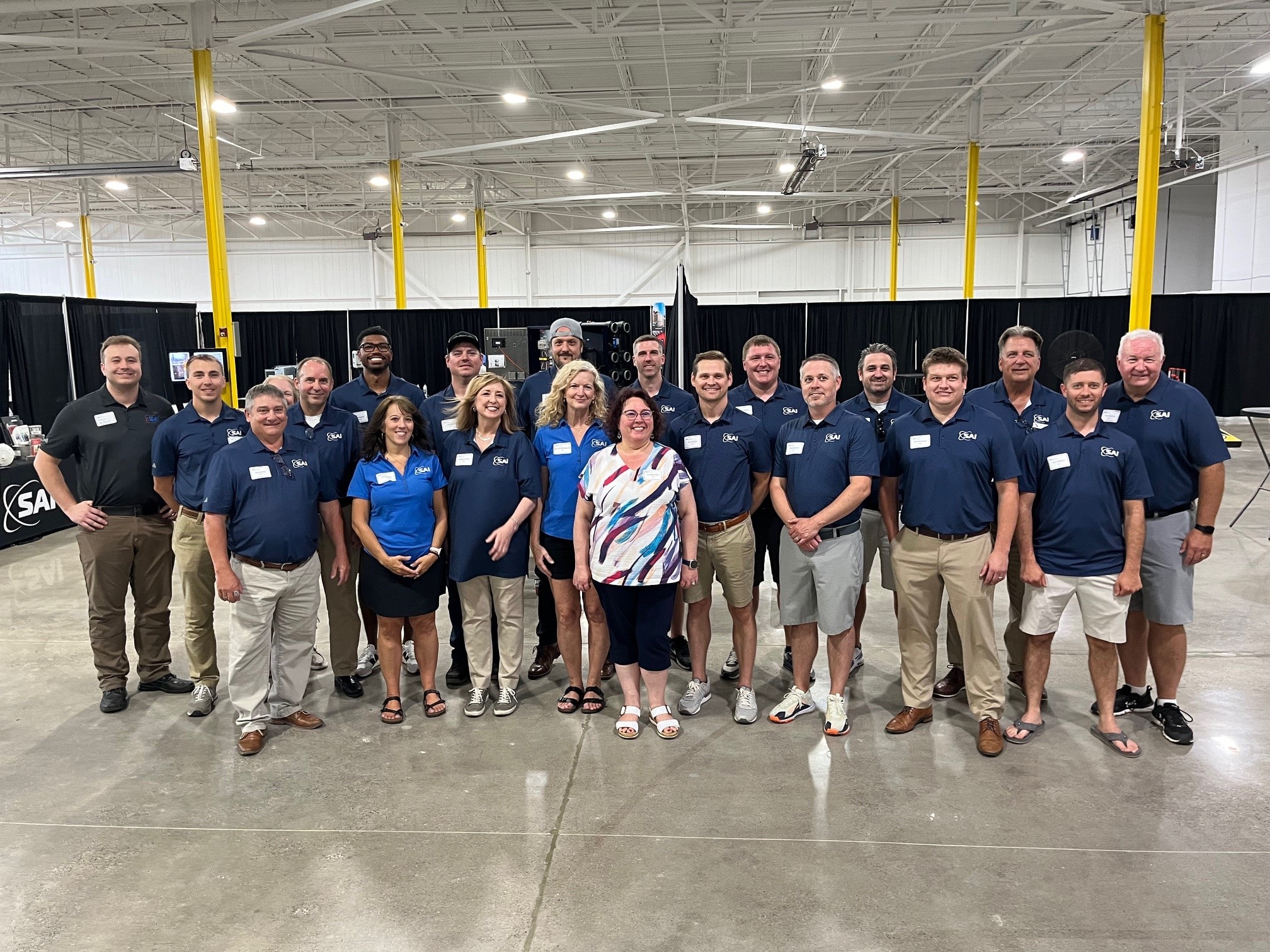 A group of people posing together in a large indoor space. They are wearing matching navy blue shirts, and some have name tags. The space has a high ceiling with visible metal beams and yellow support columns. Black curtains and various equipment are visible in the background.