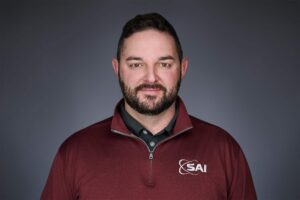 A man with short dark hair and a beard wearing a maroon sweater with 'SAI' logo and a gray polo shirt underneath, posed against a gray background.