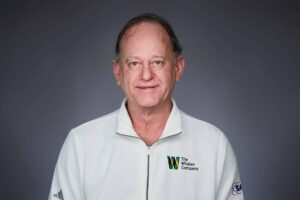 A man with short hair wearing a white jacket with a zipper and an embroidered logo for The Whalen Company on the left side. He is posing against a plain gray background, smiling slightly at the camera.