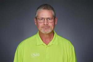A middle-aged man with short gray hair and glasses wearing a bright yellow polo shirt with the logo 'SAI' on the left side, standing against a neutral gray background.