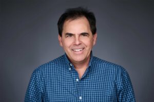 A man with dark hair smiling at the camera, wearing a blue checkered shirt, set against a plain dark background.