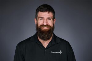 A man with a full beard and smiling expression wearing a black polo shirt with a company logo on the chest, set against a plain dark background.