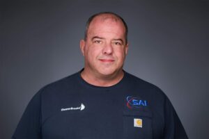Portrait of a middle-aged man with short hair wearing a dark-colored t-shirt featuring logos from CleaverBrooks, SAI, and Carhartt against a neutral gray background.