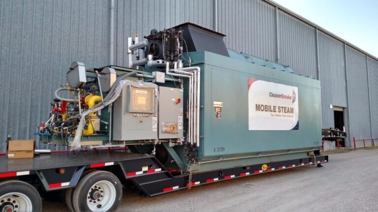 A large mobile steam boiler unit mounted on a trailer situated outside a metal industrial building. The unit, labeled 'CleaverBrooks MOBILE STEAM,' includes complex piping and control panels. The trailer has double axles and is parked on a gravel surface.