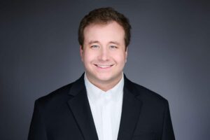 A man in a black suit and white shirt smiling against a gray background. He has short brown hair and is looking directly at the camera.
