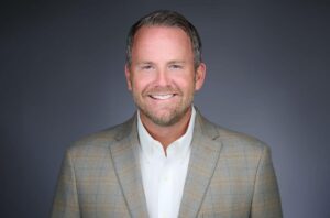 A man with short hair and a beard, wearing a light grey plaid suit jacket and a white shirt, smiling against a plain grey background.