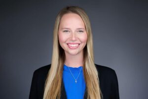 A woman with long blonde hair is smiling in a professional portrait. She is wearing a blue top and a dark blazer, with a silver necklace that features a small pendant. The background is a plain gray color.