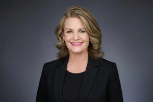 Portrait of a professional woman with blonde hair, wearing a black suit jacket, smiling against a gray background.