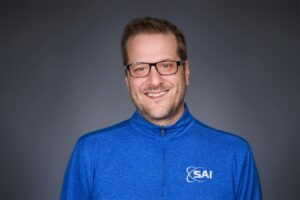 A smiling man wearing glasses and a blue jacket with the 'SAI' logo, standing against a plain dark background.