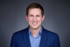 Portrait of a man wearing a blue suit jacket and a blue checked shirt, smiling at the camera against a plain gray background.