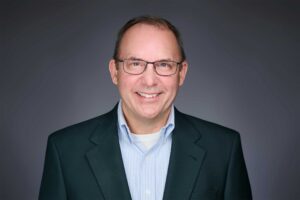 A man wearing glasses and a dark suit with a light blue striped shirt, smiling against a plain grey background.
