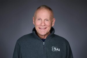 A smiling man wearing a dark gray zip-up jacket with 'SAI' logo on the left side, set against a plain gray background.
