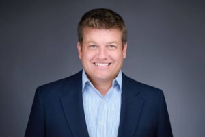 A man wearing a dark blue suit and light blue checkered shirt, smiling against a grey background.