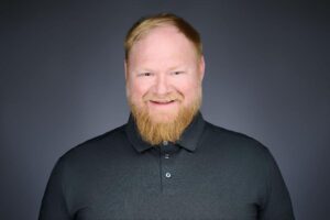 A man with a light beard and short hair, smiling at the camera. He is wearing a black collared shirt and standing against a plain, dark background.