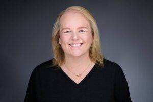 A person with light hair is smiling in a professional headshot against a dark gray background. They are wearing a black sweater and a gold necklace.