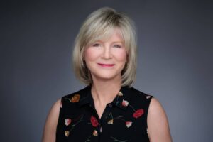 A woman with short blonde hair smiling at the camera. She is wearing a black sleeveless top adorned with colorful floral patterns. The background is a smooth gradient of dark gray, highlighting her features.
